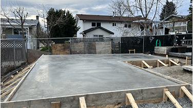 A newly poured concrete slab surrounded by wooden framing and fencing, with residential buildings in the background.