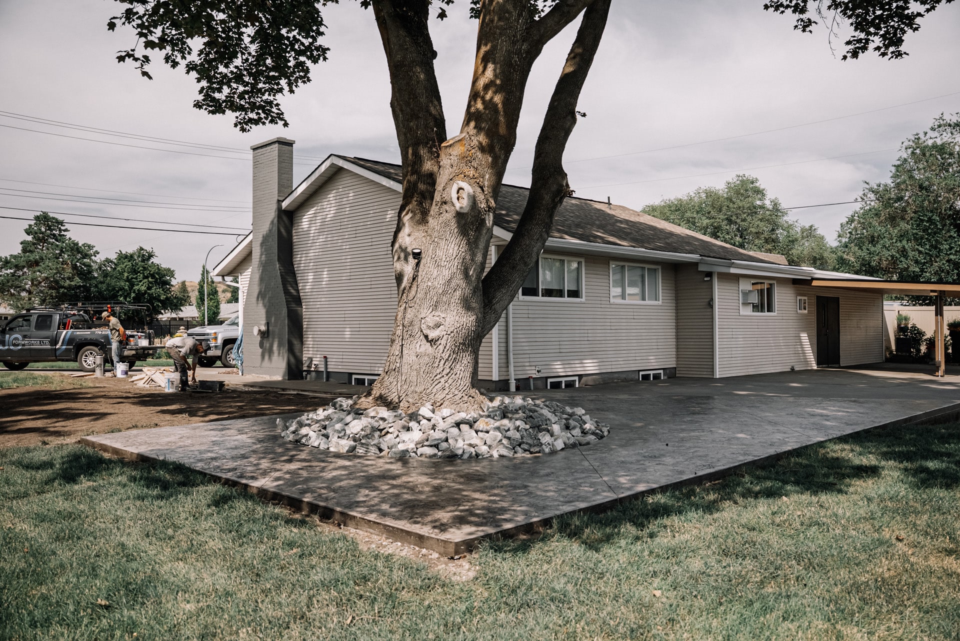 A yard with a large tree, a house with new siding, and two workers near construction materials.