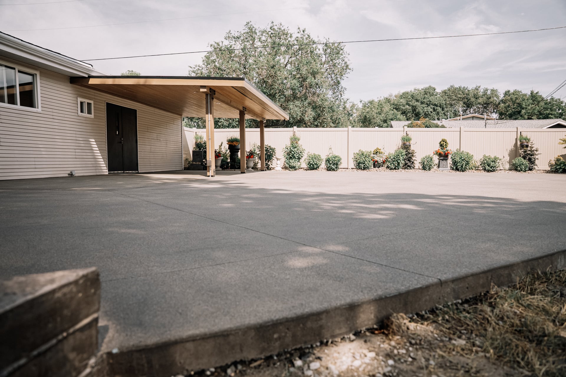 A concrete patio area in front of a house with a covered porch, surrounded by flower planters and greenery.