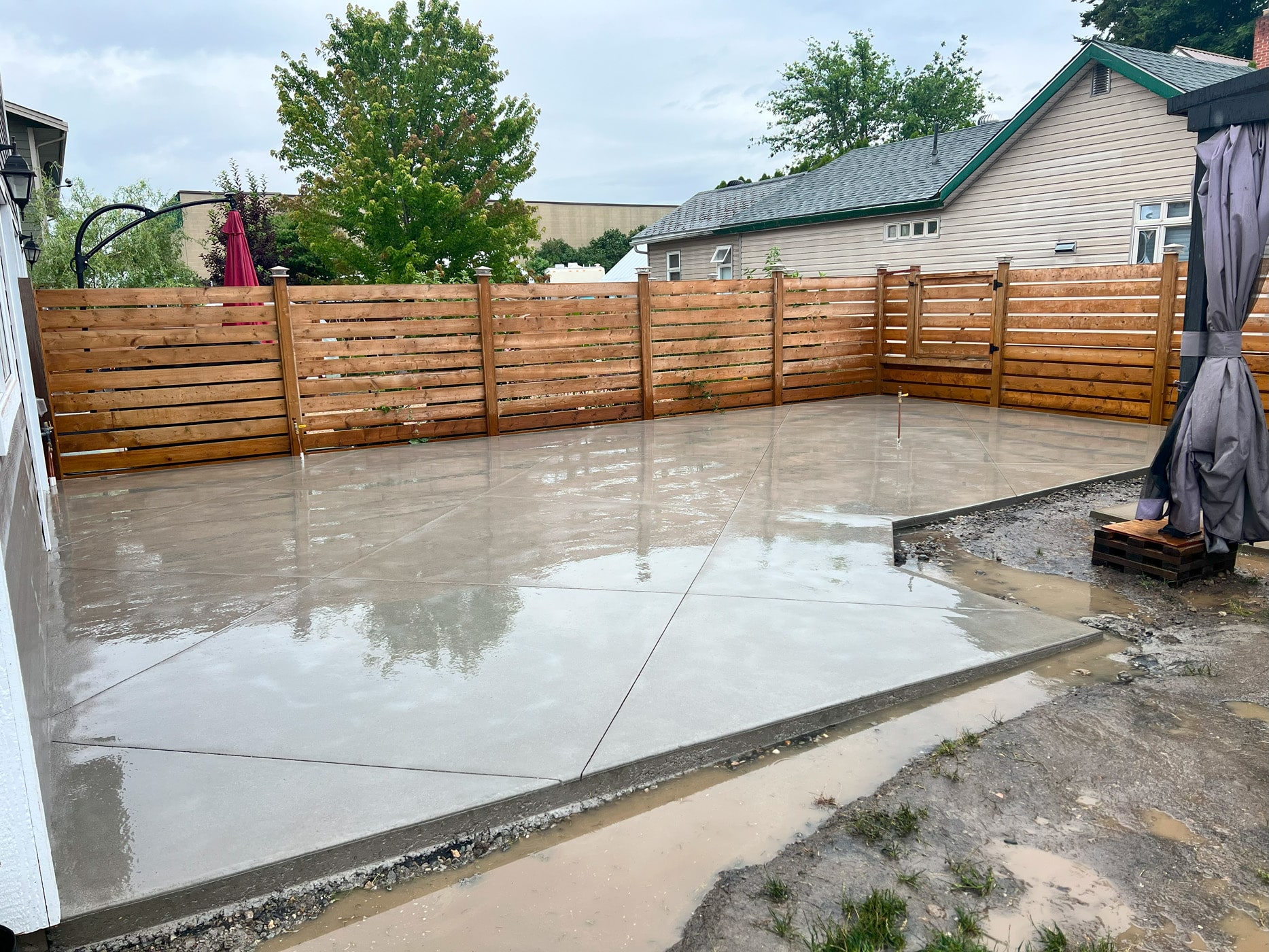 A newly installed, wet concrete patio surrounded by wooden fencing and a few trees, with a gray sky in the background.