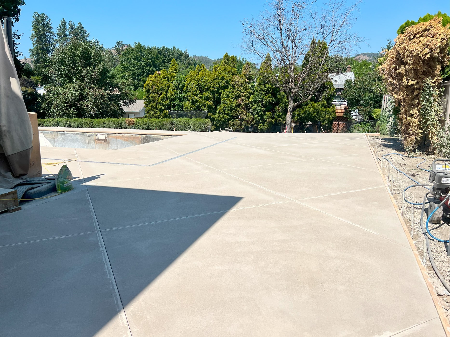 A concrete surface with a partially constructed pool area, surrounded by trees and shrubs under a clear blue sky.