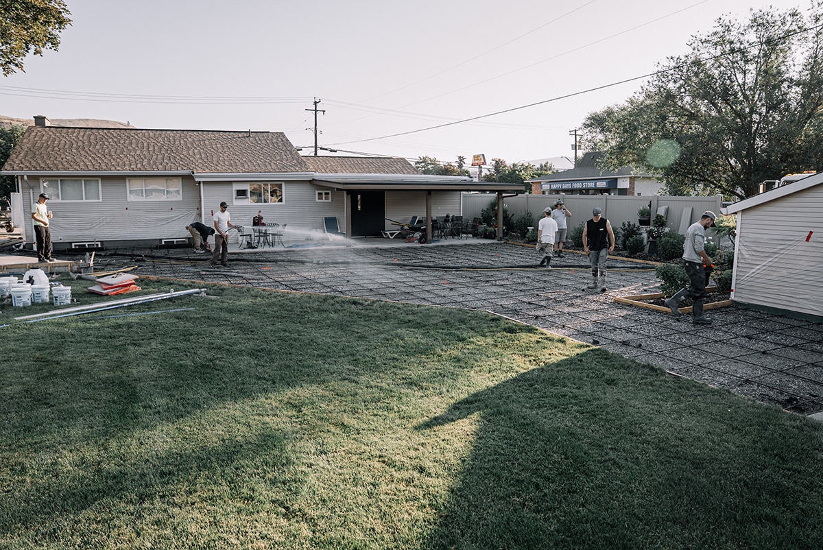A group of people working on a backyard construction project, pouring concrete on a gravel surface, with a house and a shed in the background.