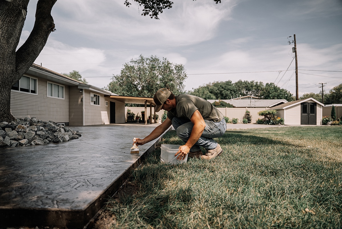 A man is applying a coat of sealant to a concrete patio while crouching on the grass near a house.